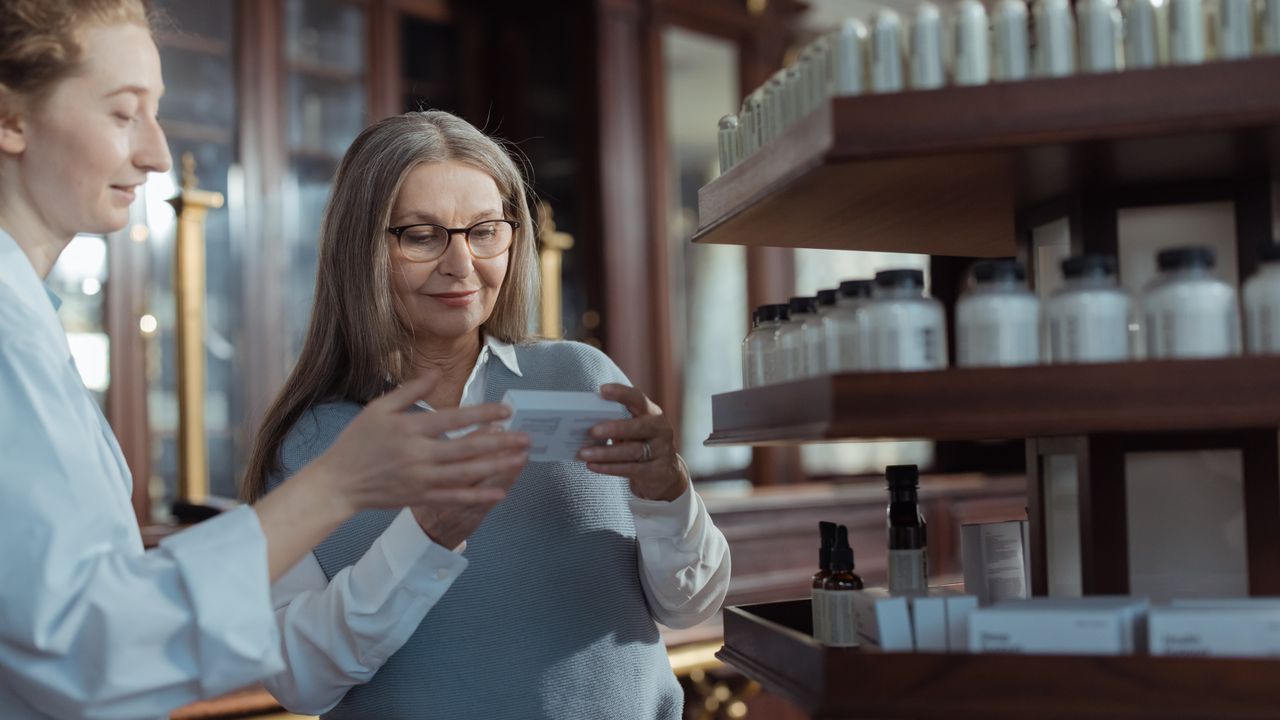 Dona de farmácia mostrando uma caixa de produto para uma cliente na loja